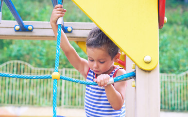 Little Caucasian boy in striped t-shirt hang the blue bar by his hand to exercise at out door playground in summer day.