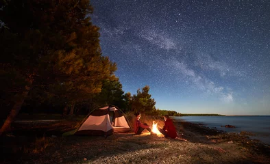 Keuken foto achterwand Kamperen Nacht kamperen aan de wal. Man en vrouw wandelaars rusten voor tent bij kampvuur onder avondhemel vol sterren en Melkweg op blauwe water en bos achtergrond. Buiten levensstijl concept