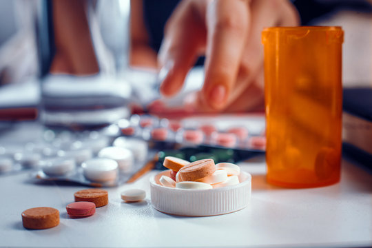 Blurred hand reaches for the orange pills laying on the table. Many other tablets laying blurred on the table near the spilled pills.