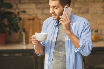 Morning news! Handsome young man drinking coffee at home in the loft kitchen, using phone.