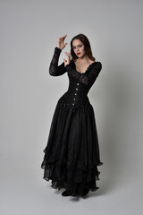 full length portrait of brunette girl wearing long black gown with corset. standing pose on grey studio background.