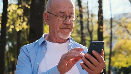 Portrait of senior man using mobile phone, holding device, texting, surfing internet, and using app. Senior man with a beard and wearing glasses looking at camera, smiling while rest in the park - Powered by Adobe