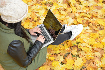 Girl in hipster with laptop in autumn park. A woman in a cap using a laptop while sitting on fallen leaves. Freelancer in the hat uses remote communication technology. Remote work. View from above