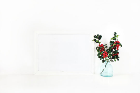 Horizontal white blank wooden frame mockup. Holly berry branches in blue glass vase on white table. Styled stock feminine photography. Home decor. Christmas winter concept.