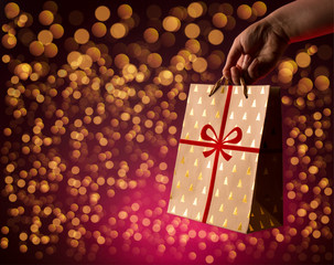 Woman holding glittering paper shopping bag with ribbon on bokeh glittering red background