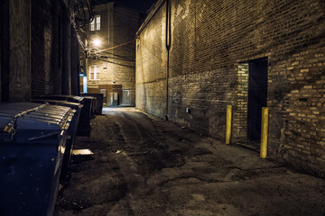 Dark and scary downtown urban city street corner alley with an eerie vintage industrial warehouse factory entrance and dirty dumpsters at night