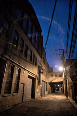 Dark and scary downtown urban city street alley scene with an eerie vintage industrial warehouse factory skyway at night