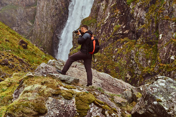 Nature photographer tourist with camera shoots while standing on the mountain against a waterfall.