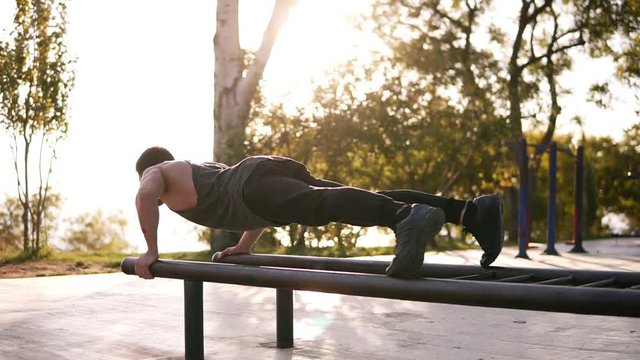 Athletic young caucasian man doing push ups on parallel bars in local outdoors gym in the park. Man wearing black sports clothes