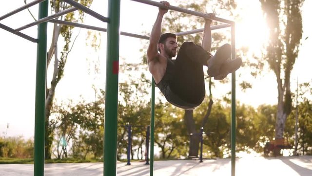 Fitness, sport, training and people concept - strong bearded man doing legs exercises while hanging on the horizontal bar in the park. Abdominal and legs exercise in outdoors gym