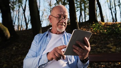 Old man is using tablet computer in the park. Senior man with a beard and wearing glasses, sits on a park bench and uses hir portrable computer - Powered by Adobe
