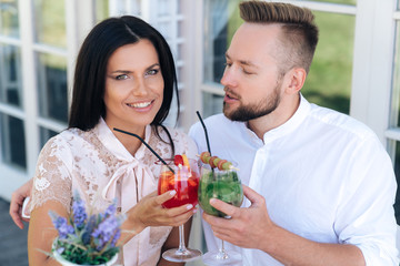 A close-up, a radiant young woman smiling broadly, showing her even white teeth, sitting at a table with her boyfriend, having a good time, drinking a cocktail. the man wants to kiss the girl.