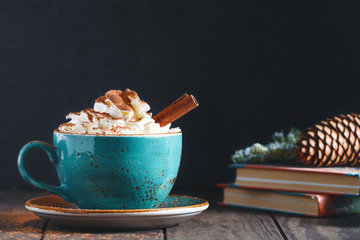 Hot chocolate with cream and cinnamon stick in a blue ceramic cup on a table with a books. The...
