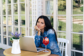 The frustrated girl sits with a displeased look in the cafe, looks thoughtfully away, tensely thinks. The model sits with a laptop at a table in a cafe.