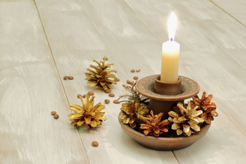 Ceramic candlestick and burning white wax candle, multicolored pine cones and coffee grains on light wooden background.
