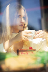 Beautiful blonde model spending time in the restaurant with a cup of coffee. Shot through window glass
