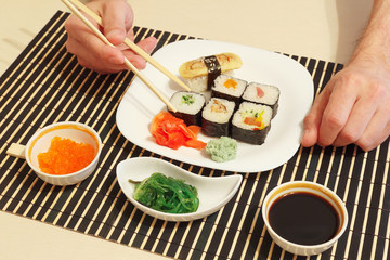 Hands taking sushi with chopsticks. Sushi set, hiyashi wakame salad, soy sauce and salmon caviar on a black bamboo mat.