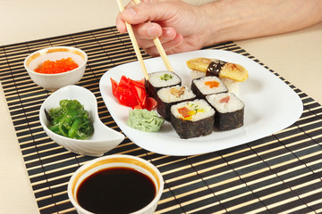 Hands taking sushi with chopsticks. Assorted sushi, chuka salad, soy sauce and salmon caviar on a black bamboo mat.