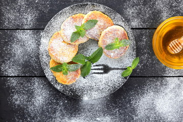 Cottage cheese pancakes with fresh mint sprinkled with powdered sugar. Syrniki on dark wooden boards. Healthy food for breakfast. Cottage cheese pancakes and honey on wooden stand