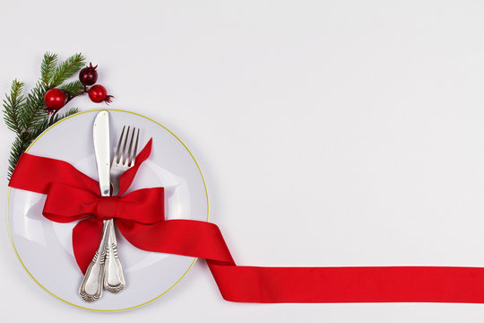 Christmas composition with plate, cutlery, pine branches, ribbon and red berries on white table. Winter holidays and festive background. Christmas eve dinner, New Year food lunch.  top view
