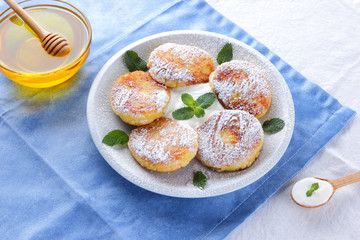 Cottage cheese pancakes sprinkled powdered sugar and fresh mint. Cheesecakes on white plate. Sour cream in wooden spoon. Honey and dairy products on white blue background. Healthy food