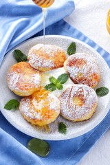 Cottage cheese pancakes sprinkled powdered sugar and fresh mint. Cheesecakes on white plate. Sour cream in wooden spoon. Honey and dairy products on white blue background. Healthy food
