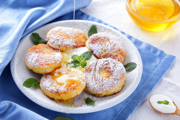 Cottage cheese pancakes sprinkled powdered sugar and fresh mint. Cheesecakes on white plate. Sour cream in wooden spoon. Honey and dairy products on white blue background. Healthy food