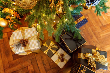 Fototapeta na wymiar Classic Christmas decorated New year gold paper gift box with golden ribbon bow near Christmas tree. Modern dark classical style interior design apartment. Christmas eve at home