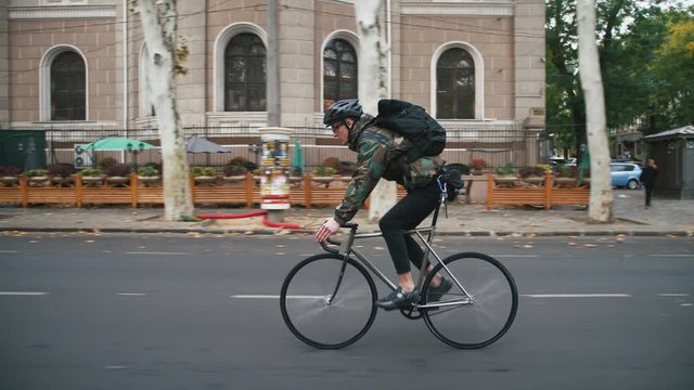 Young hipster man in helmet riding fixed gear bicycle in the city during autumn morning, side view