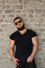 Fototapeta premium Bodybuilder in sunglasses portrait. Muscular man with a beard in a tight T-shirt is standing against a brick wall. Stylish and sporty young man.