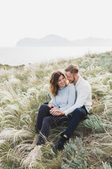 Happy young loving couple sitting in feather grass meadow, laughing and hugging, casual style sweater and jeans - 231612675