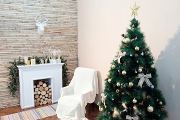 Christmas tree stands near the bedside table and fireplace against the white wall. Theme happy new year. Paper deer head hanging on the wall of wooden planks.