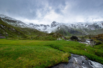 Schnee auf Berggipfeln der Alpen mit einen Fluß und grüner Wiese im Vordergrund