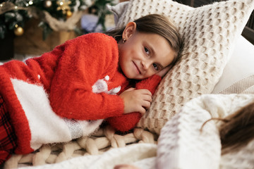 Close up portrait of young caucasian child girl lying on the pillow at cozy bedroom. Christmas tree on the background. Girl smiling, enjoying holidays, Merry Christmas, New year.