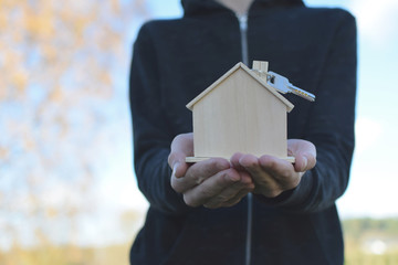 Young man holding and giving wooden house model with key outdoors on blurred landscape background. Real estate, buying moving new home or renting property or apartments concept.