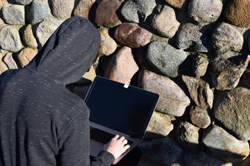 Young man wearing hoodie hacking laptop with black screen trying to crack password back view outdoors on decorative stone wall background. 