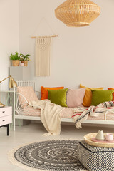 Rattan chandelier above single metal bed with pastel pink, olive green, orange and yellow pillows, patterned carpet and pouf on the floor, macrame on the white wall with copy space