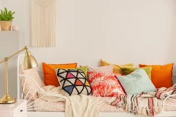 Fotobehang Boho Patterned and colorful pillows on comfortable white bed in stylish bedroom interior with golden lamp on bedside table and handmade macrame on white wall, real photo with copy space  © Photographee.eu
