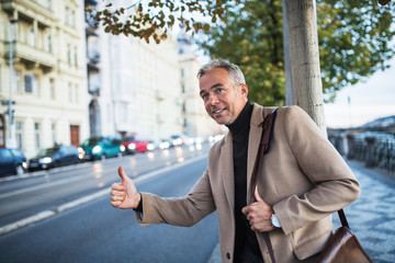 Mature businessman standing on the street in city, raising his hand to hail a taxi cab.