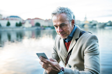 Mature businessman with smartphone standing by river Vltava in Prague city.