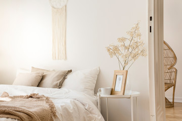 Bedside table with mug and framed picture next to comfortable king size bed with linen bedding and beige blanket, real photo