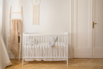 Scandinavian nursery with white wooden crib and macrame on the wall in tenement house, real photo...