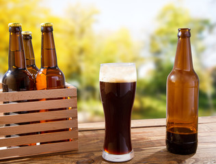 cup of dark beer and bottles on wooden box on table on blurred park background