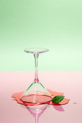 Glass martini glass and spilled liquid on a pink-green background. Modern art photography.