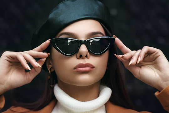 Outdoor close up fashion portrait of young beautiful woman wearing black cat eye sunglasses, leopard print hoop earrings, leather beret, white turtleneck. Model looking up
