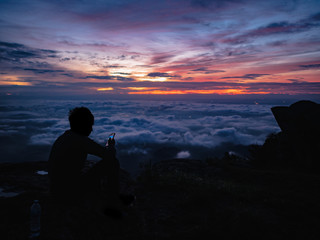 Silhouette People Sit and Look at his phone on the cliff with beautiful sunrise sky on on Khao Luang mountain in Ramkhamhaeng National Park,Sukhothai province Thailand