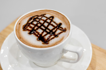 The white cup of hot capuchino and chocolate syrup on top on the wooden table background.