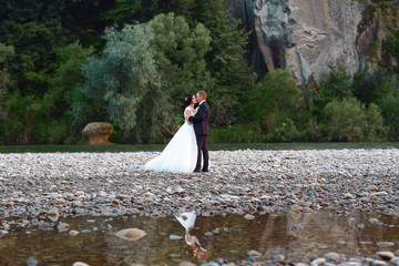Romantic wedding moment of newlywed couple hugging near blue mountain river, sensual groom embracing gorgeous bride from behind river. Wedding day for man and woman