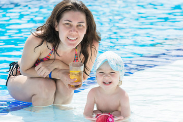 Beautiful mother with cocktail in her hands and small child are sitting in blue pool with smile on her face.