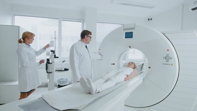In Medical Laboratory Female Radiologist and Male Doctor Control and Monitor MRI or CT Scan with Female Patient Undergoing Procedure. High-Tech Modern Medical Equipment. 